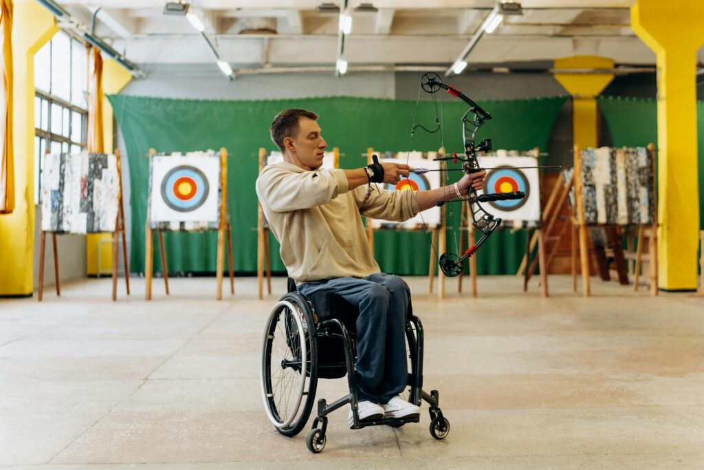Focused young man in a wheelchair practicing archery indoors, aiming with determination at a target.