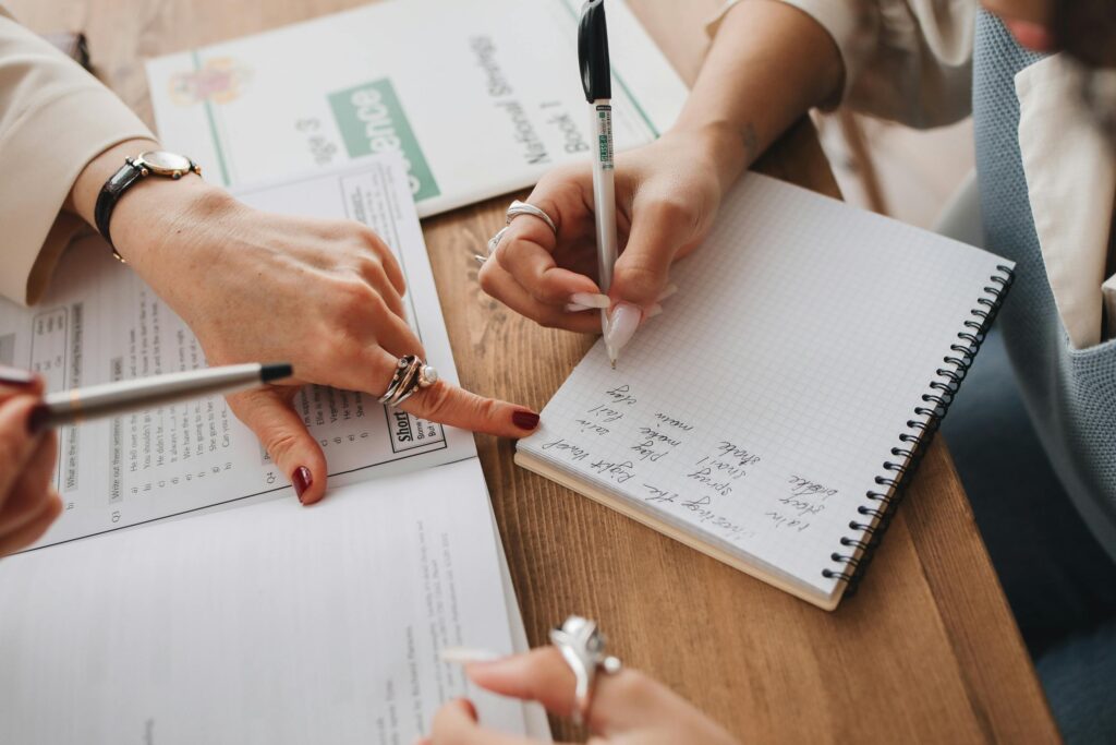 Close-up of hands writing in a notebook while studying documents on a wooden table.